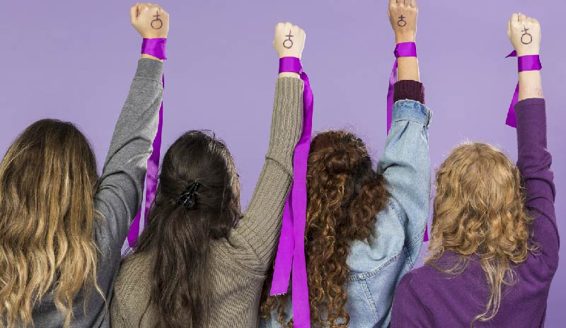 Cuatro mujeres de espalda con su mano derecha arriba sobre la cual tienen el símbolo de mujer y una cinta morada.