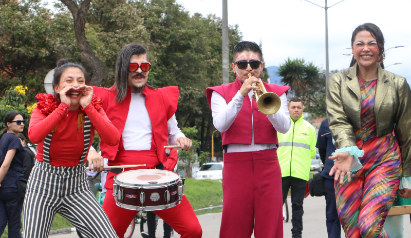 Cuatro artistas en personajes. De izquierda a derecha: mujer joven, con sus manos en la boca emulando un megáfono. Tiene pantalón a rayas verticales negras y blancas con tirantas. Su camisa es roja con flequillo superior. Su cabello es negro y se encuentra recogido. A su lado derecho, se encuentra un hombre jhoven con una redoblante de cabello hasta los hombros que está todo hacia el lado izquierdo, gafas rojas, camisa de manga blanca, chaleco rojo con hombreras puntudas que sobrepasan los brazos.