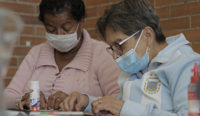 Dos mujeres mayores, al lado izquierdo una con una blusa rosada y otra con una chaqueta blanca al lado derecho, construyendo manualidades 