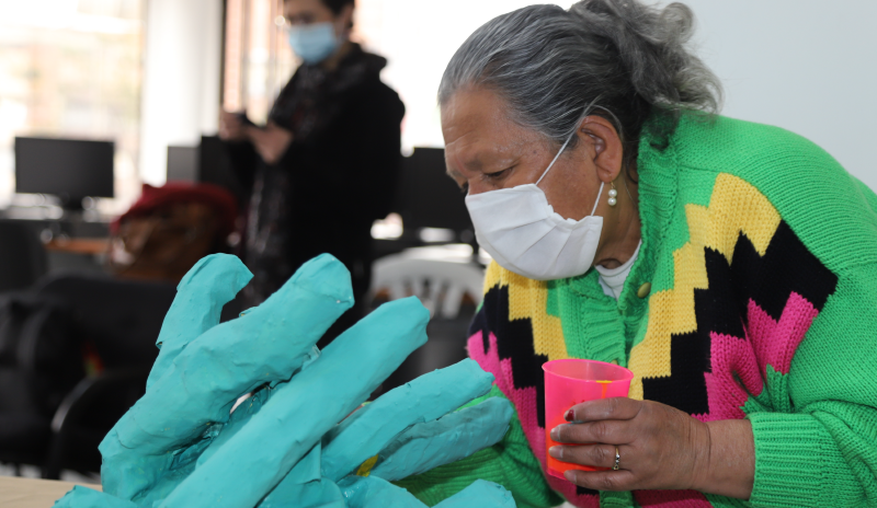 Mujer pintando una escultura de papel mache con color verde pastel. Usa un saco en color verde mente con una figura en V amarilla, negra y fuscia. Usa tapaboca, de tex blanca y con cabello gris y blanco. 