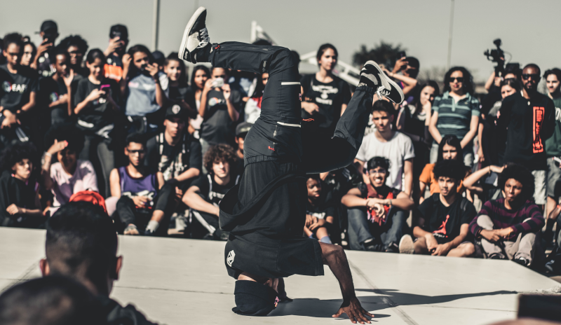 Hombre bailando hip hop. Vestido con camiseta y pantalón negro. no se alcanza a ver su rostro. Esta haciendo una figura de baile. Su cabeza se encuentra sobre el piso y los pies se encuentran en aire de manera vertical con su cara. Sus pies se encuentras distanciados uno del otro por lo que, en ambos cadsos sus rodillas se encuentran flexionadas. El bailarín se encuentra en medio de circulo formado por sus espectadores. 
