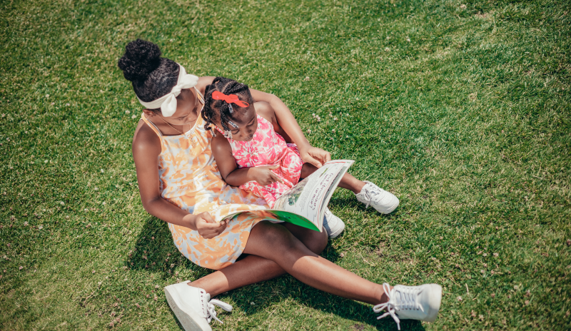 Pareja de niñas afro leyendo un libro al aire libre. No se identifican los rostros de las niñas y la imagen es tomada desde arriba. La niña más grande tiene un vestido beige y tennis blancos. La más pequeña que se encuentra entre los brazos de la mayor tiene un vestido rosado. Ambas tienen el cabello recogido.