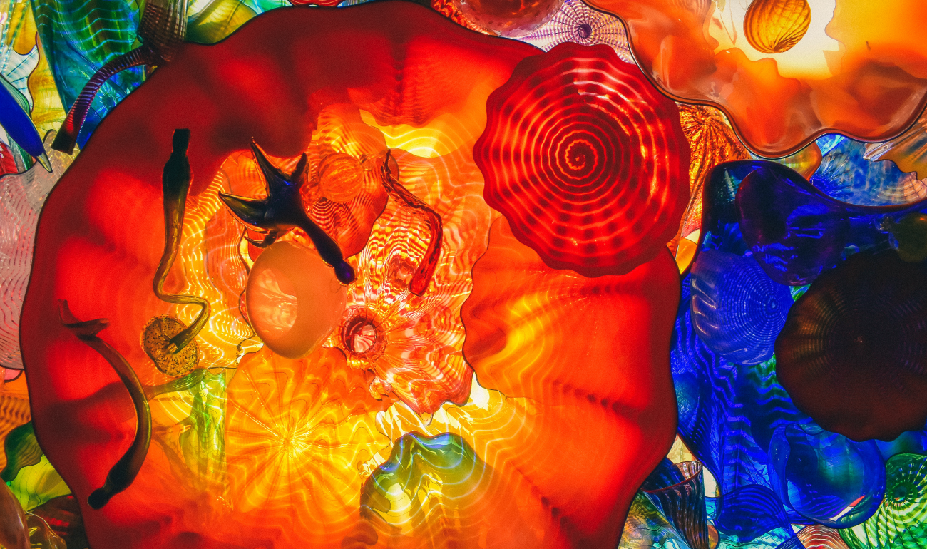 Fotografía superior de varias figuras de vidrio. La más sobresaliente es la que se encuentra del medio a la derecha de la fografía de color naranaa seguída al aldo opuesto de una de color azul. Se evidencia una mezcla de colores.