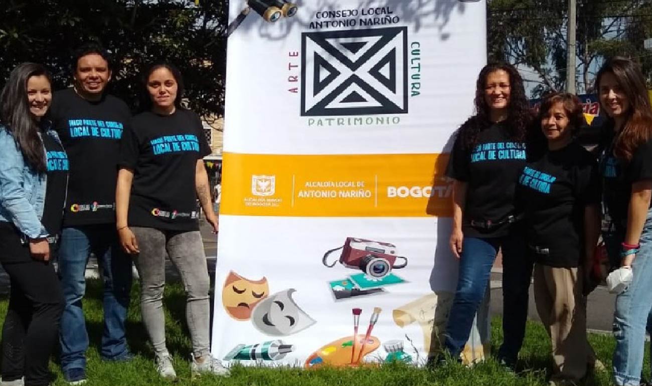 Fotografía de integrantes del Consejo Local de Arte, Cultura y Patrimonio de Antonio Nariño. Al lado izquierdo hay dos mujeres y un hombre intercalados. Ambas mujeres tienen cabello debajo del hombro en color castaño y negro respectivamente. Y al lado derecho hasy tres mujeres. Dos con cabello debajo del hombro y la mujer del medio, la mayor, tiene cabello sobre el hombre. Los seis tienen camisa negra con el texto "hago parte del consejo de cultura". En medio se encuentra el pendeón de la instancia. 