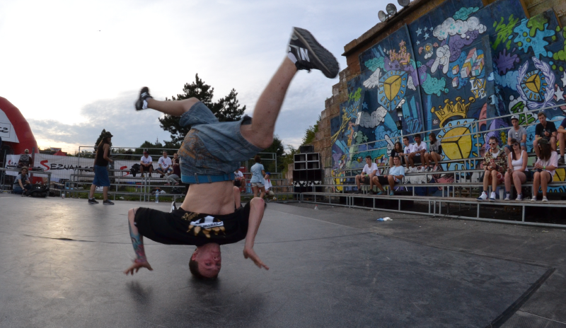 Joven haciendo una figura de hip hop. Tiene jeans, camiseta negra. Su cabeza se encuentra sobre el piso de linolio y sus pies en el aire.