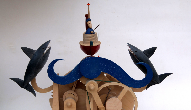 Juguete de madera, autónomata. Cuenta en el medio con un capitán de un barco, que se encuentra en el mastil y tiene uniforme azul y dos ballenas, una a cada extremo, que en el momento de mover las paláncas del juguete se mueven. 