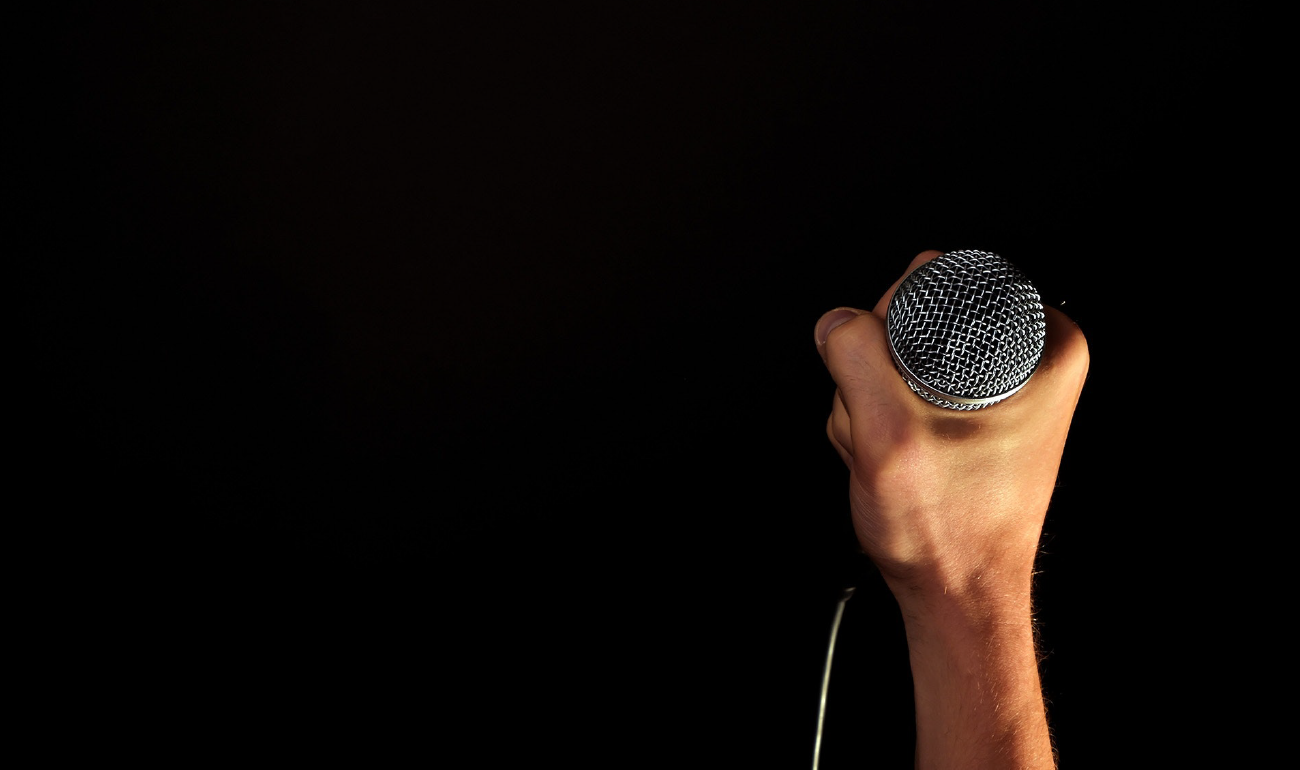 Mano con un micrófono. Esta imagen se encuentra ubicada al lado derecho de la pieza sobre fondo negro. 