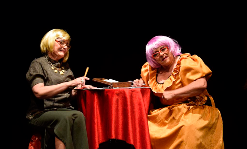 Dos artistas disfrazados de personas mayores sentadas en una mesa