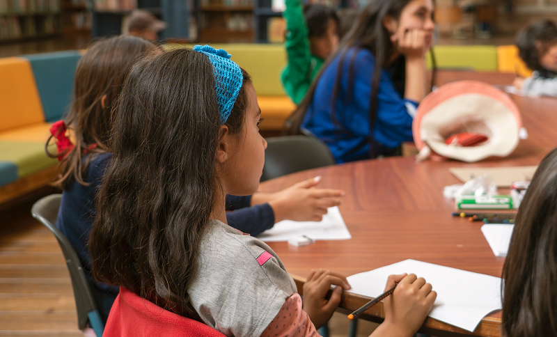 Niños y niñas alrededor de una mesa escuchando a una persona